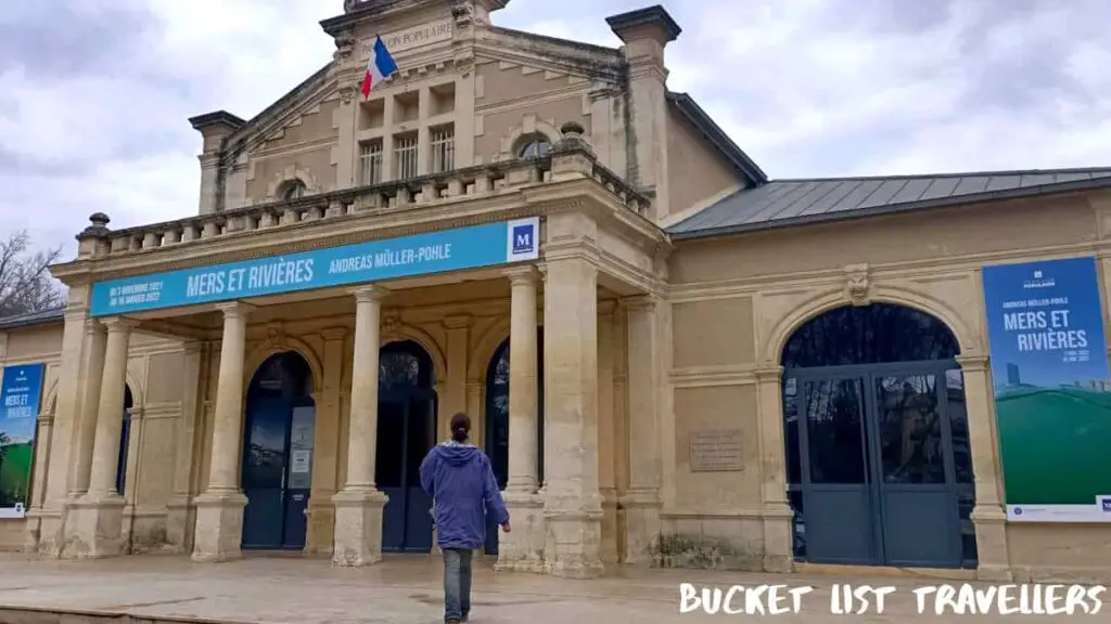 Woman wearing purple jacket and jeans walking towards Pavillon Populaire Montpellier France, signs displaying exhibition Mers et Rivieres, sandstone building with French flag