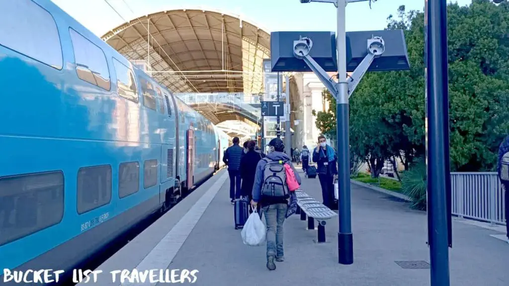 Gare de Nice-Ville - France Train Station Platform, people walking along platform beside blue train, woman wearing jean, purple coat and hat carrying backpack