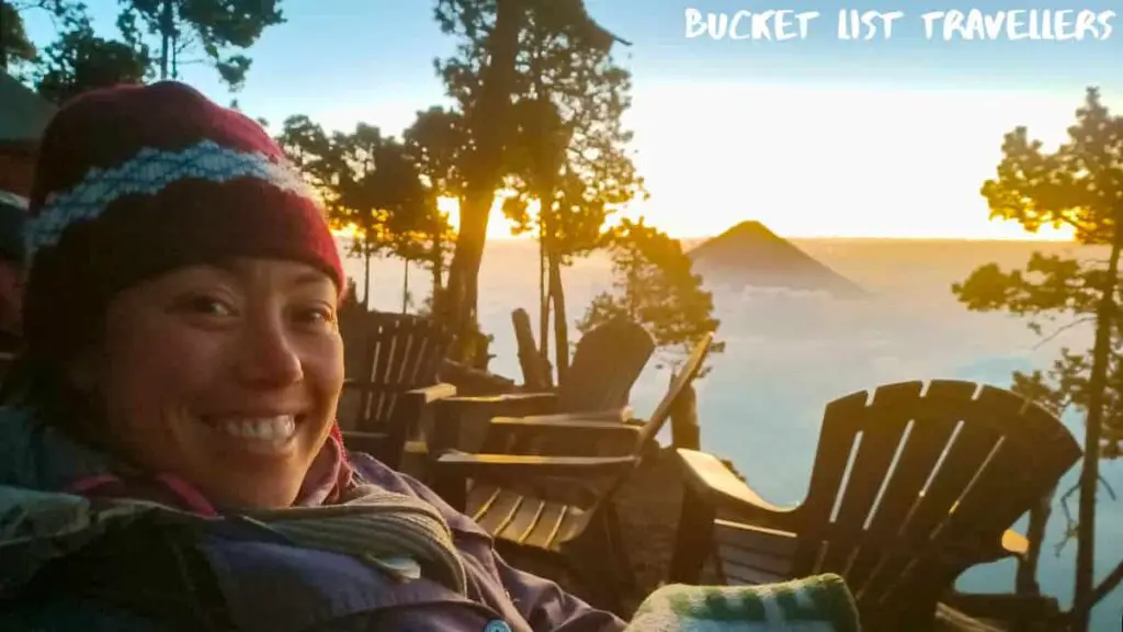 Woman wearing beanie and jacket sitting at Wicho & Charlie's campground on Acatenango Volcano Guatemala, Pacaya Volcano in the background, sunrise on a volcano, volcano surrounded by clouds