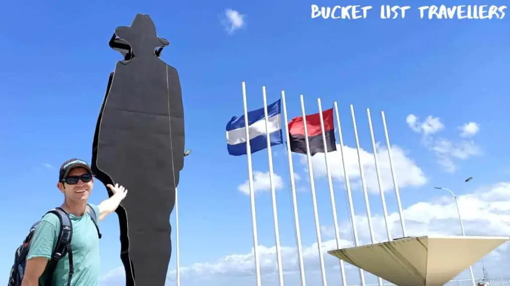 Man in green t-shirt pointing to Sandino Statue at Loma de Tiscapa Managua Nicaragua; Nicaragua and FSLN flags in the background; blue sky wtih white clouds